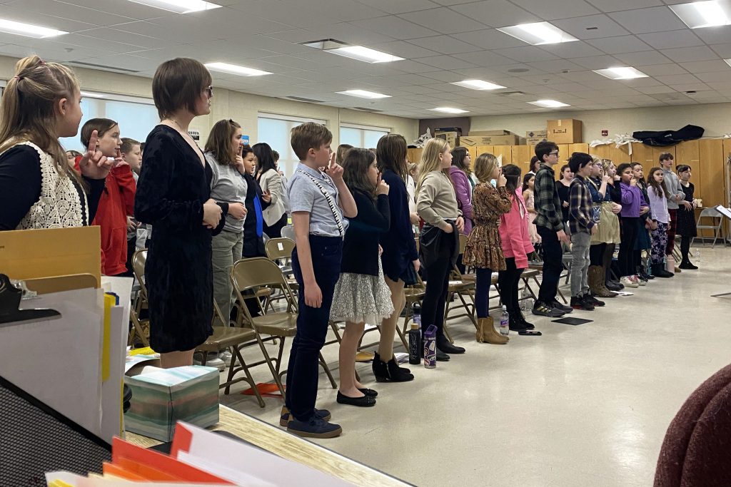 Choir students stand in class, some with their index finger propped to their lips in a "shush" gesture