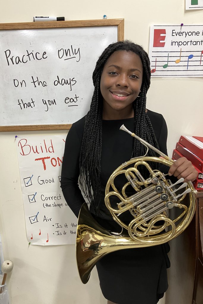 A student poses with her French Horn next to a sign that jokingly says "practice only on the days that you eat"