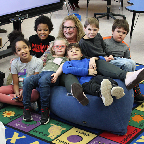 Jackie Whitman smiles while sitting on a bean bag with 6 young children nestled around her