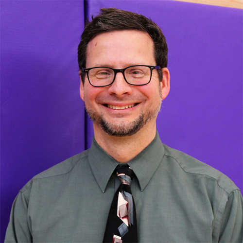 Sean Russo in shirt and tie smiles happily in front of a purple gym wall