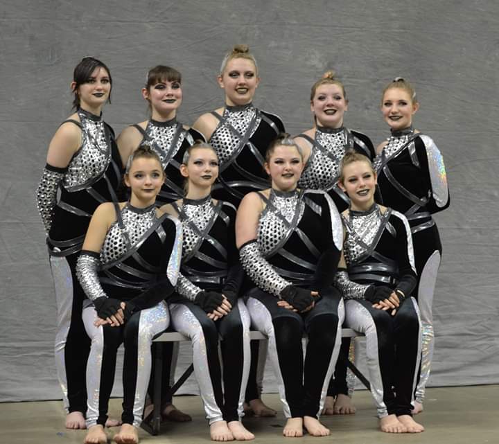 Nine members of the Amethyst Gold varsity guard team dressed in silver and black patterns stand or sit in two rows