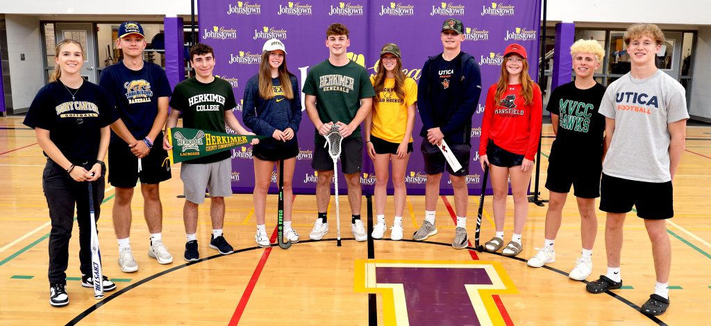 Graduating students stand in a semi circle, wearing the t-shirts of the colleges they plan to play sports at