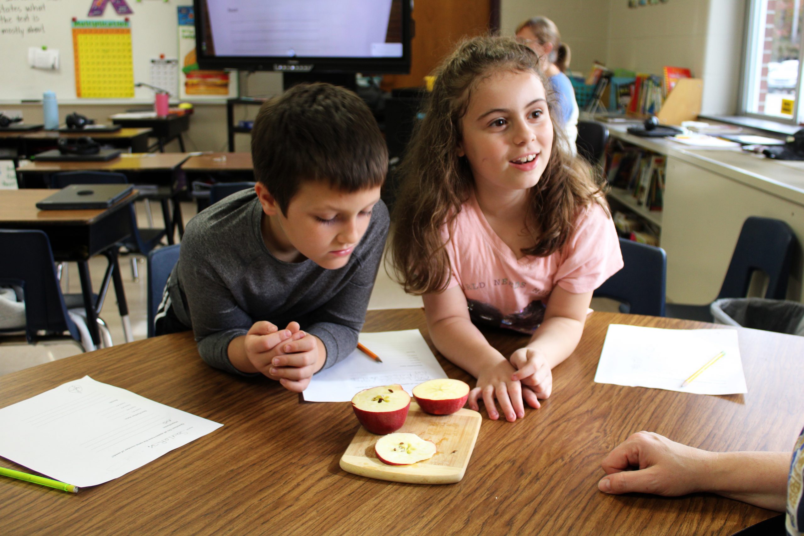 A student looks excitedly at her teacher while another studies a split apple in front of him