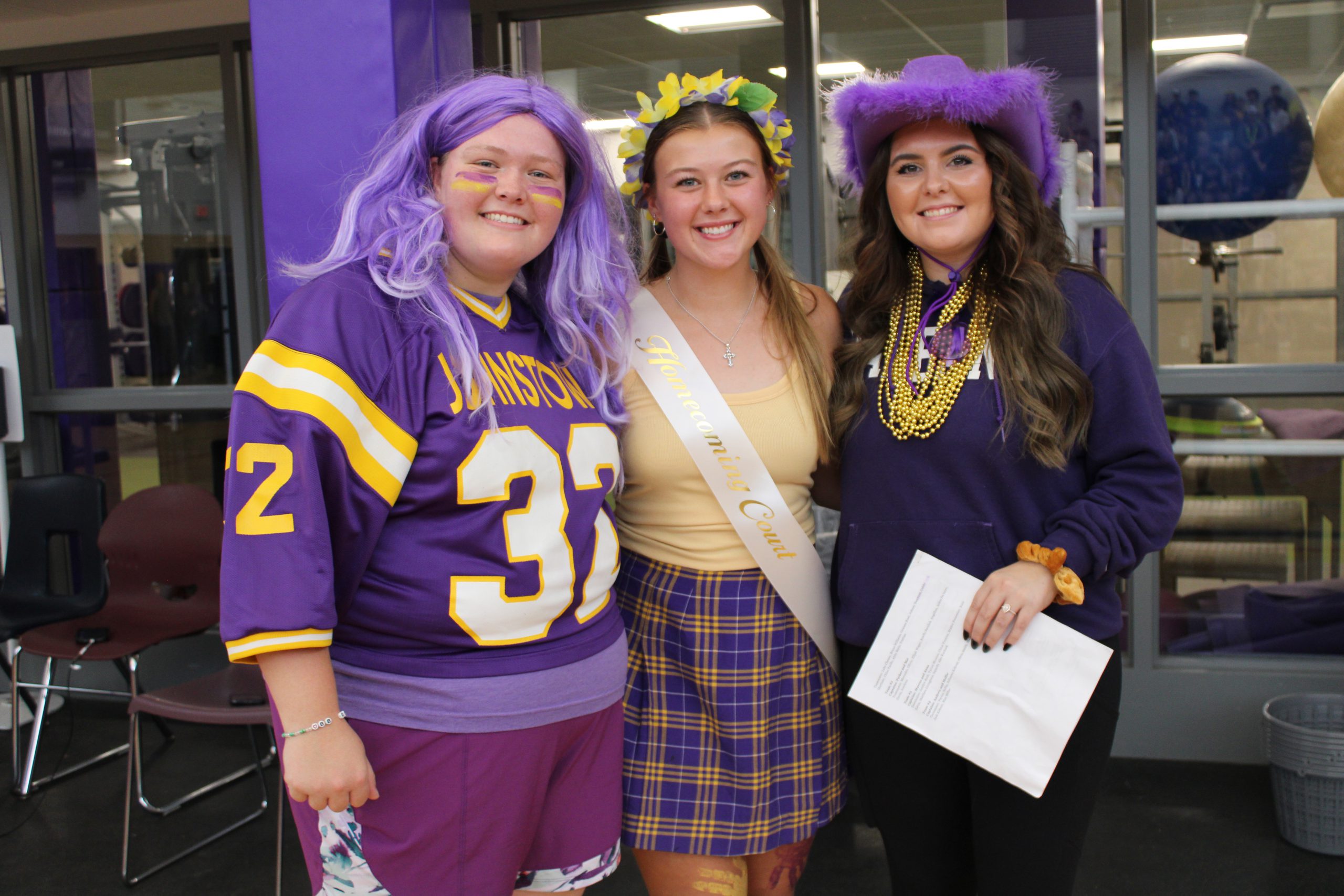 Two students gather on either side of a student wearing a sash, all three dressed in purple and gold