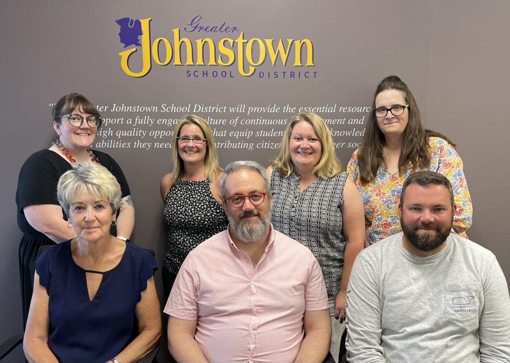 Seven members of the Board of Education gather together in front of a wall displaying the school district mission statement