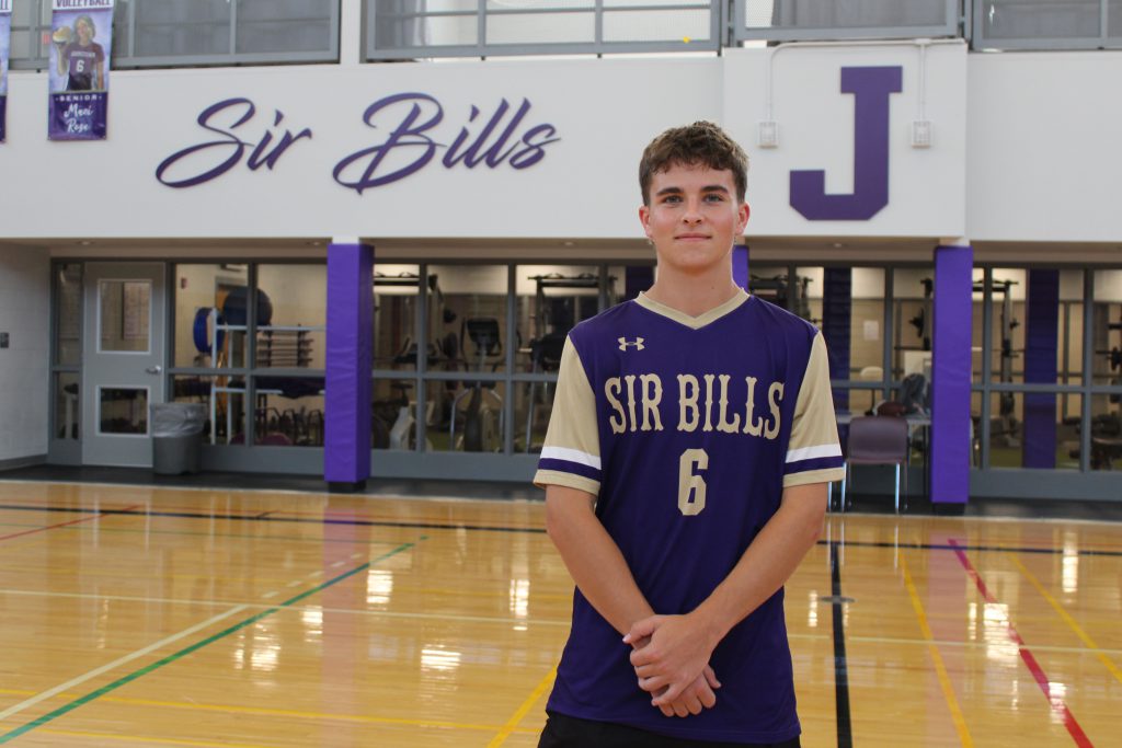 a young man wearing a Sir Bills athletic uniform poses inside of a gymnasium