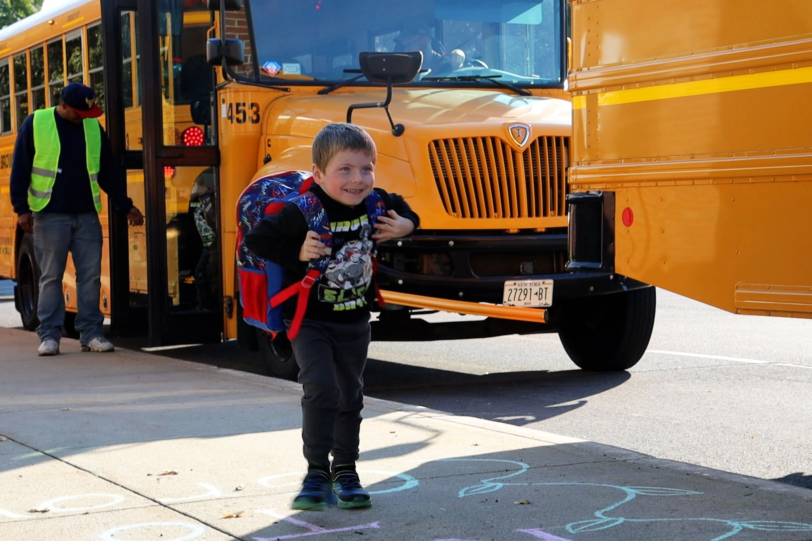 a young boy with a backack on walks on the sidewalk with a school bus behind him