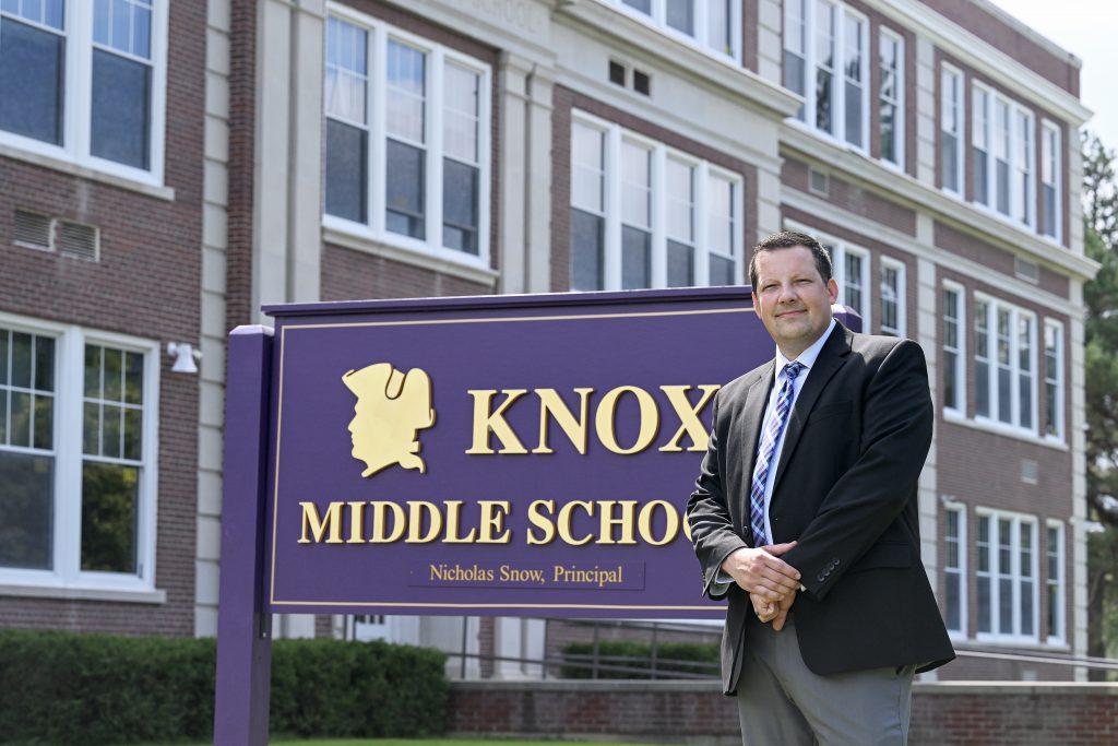 a man wearing a suit and tie stands in front of a school building and a sign that reads Knox Middle School