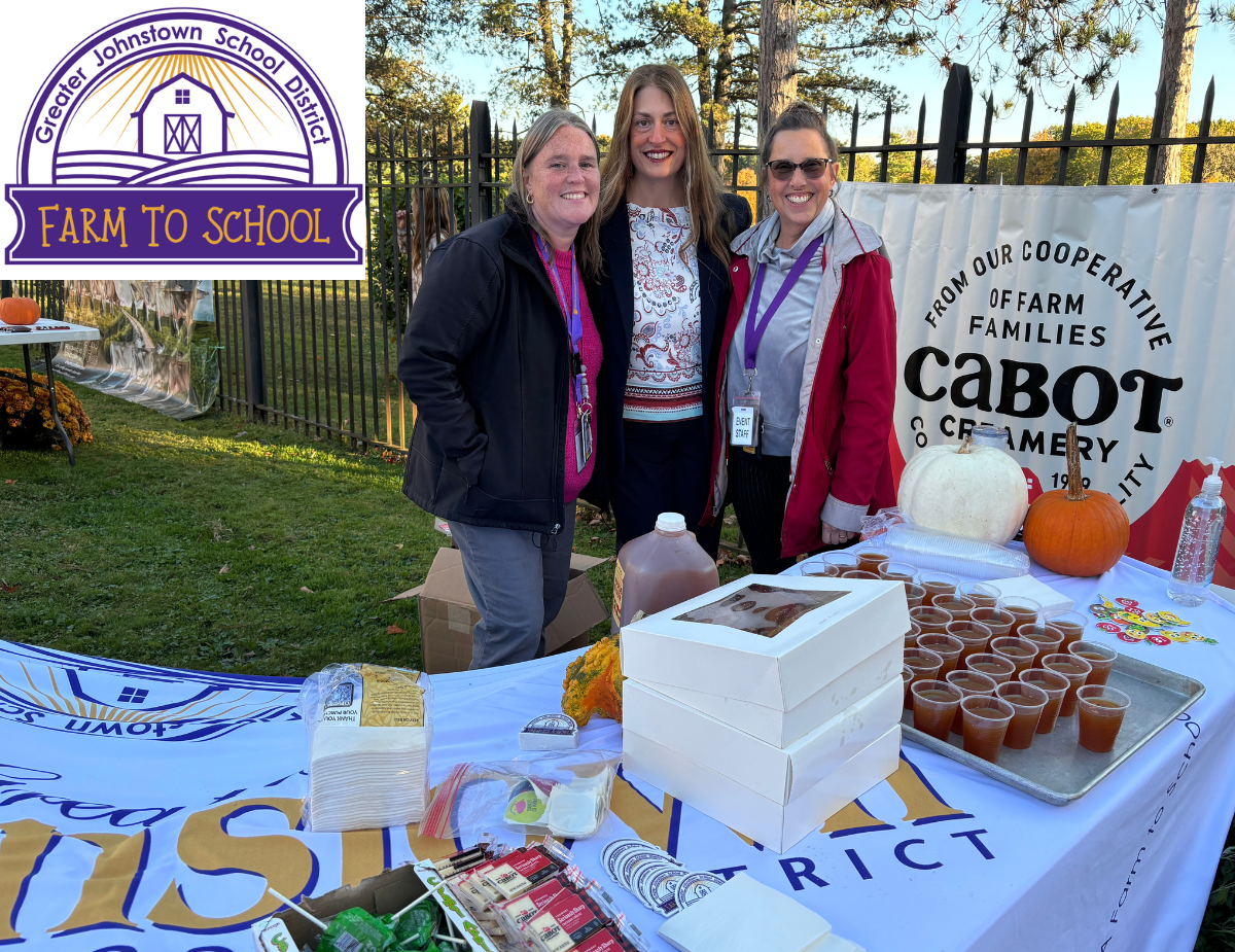 three adult women stand behind a table full of donuts and other fresh food items