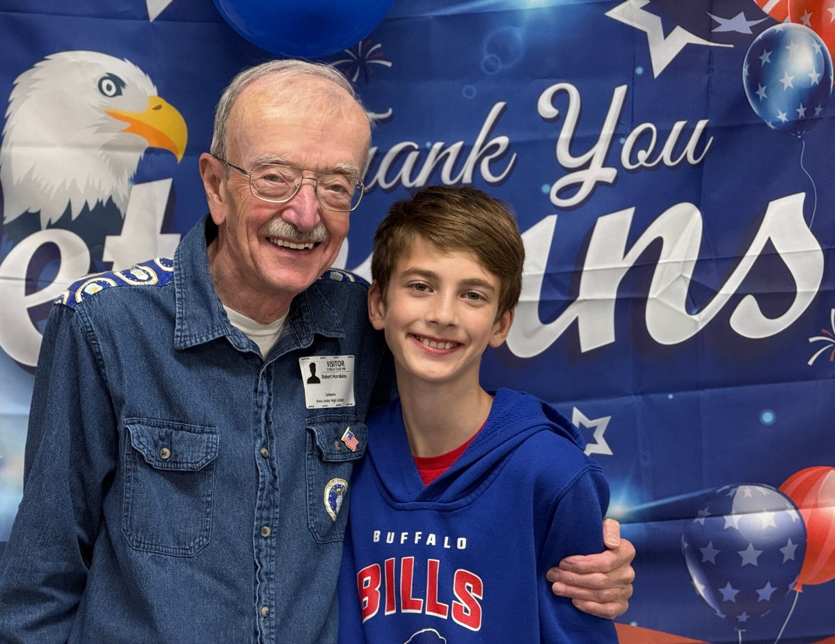 an adult male poses next to a young boy in front of a veterans day banner