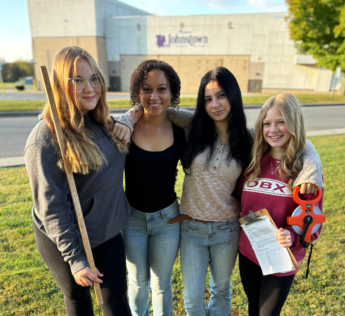 four teenaged girls stand in front of the school holding measuring sticks to be used in a lab 