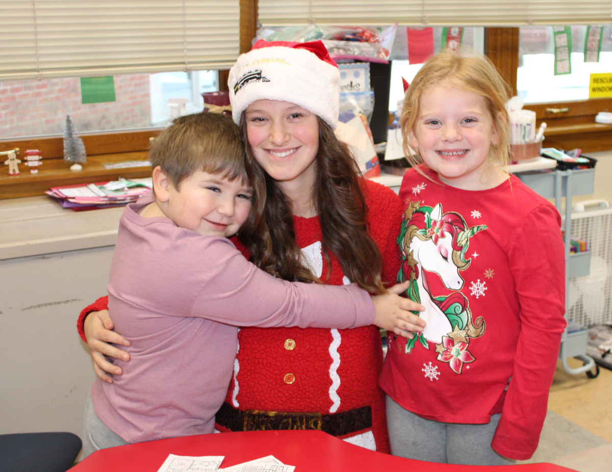 a young teen girl poses in a santa hat with two elementary students