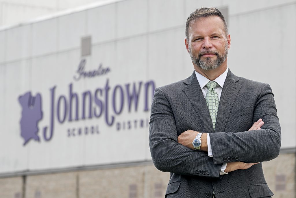 a man in a suit stands with his hands crossed in front of a school building 