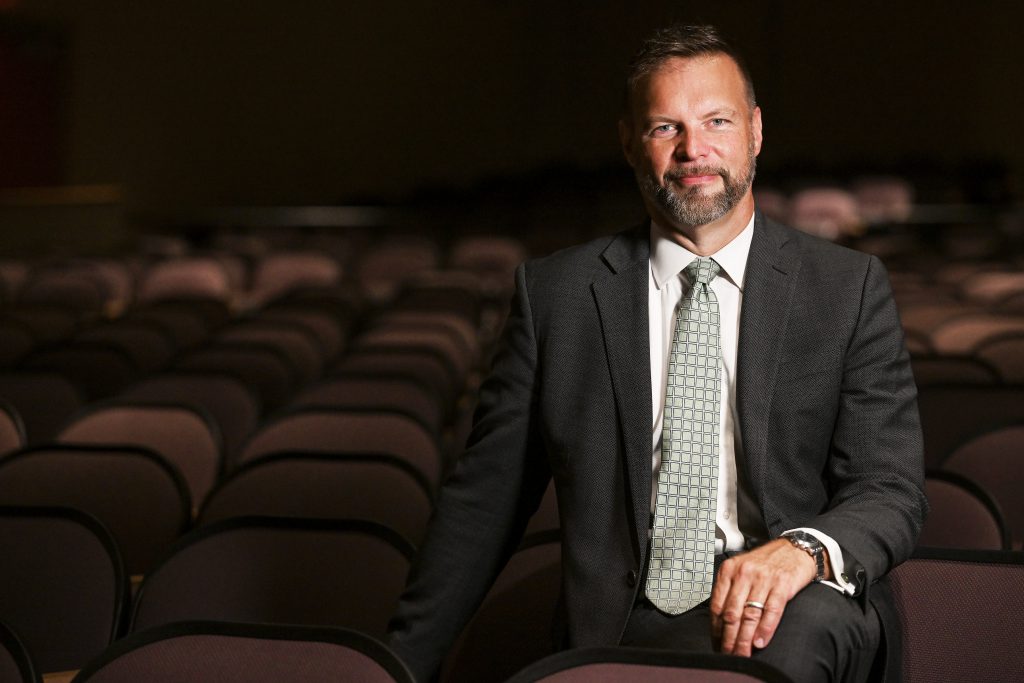 a man wearing a suit and tie sits in an auditorium