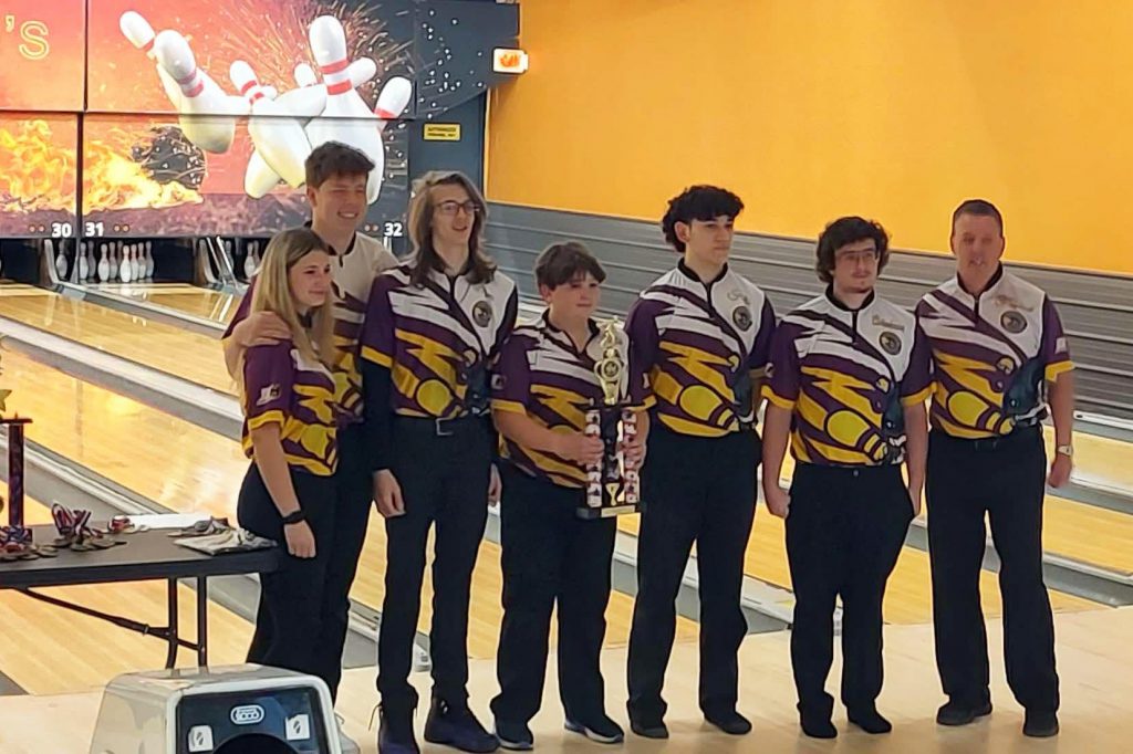 a group of 6 students and one adult pose in team jerseys on a bowling lane holding a trophy.