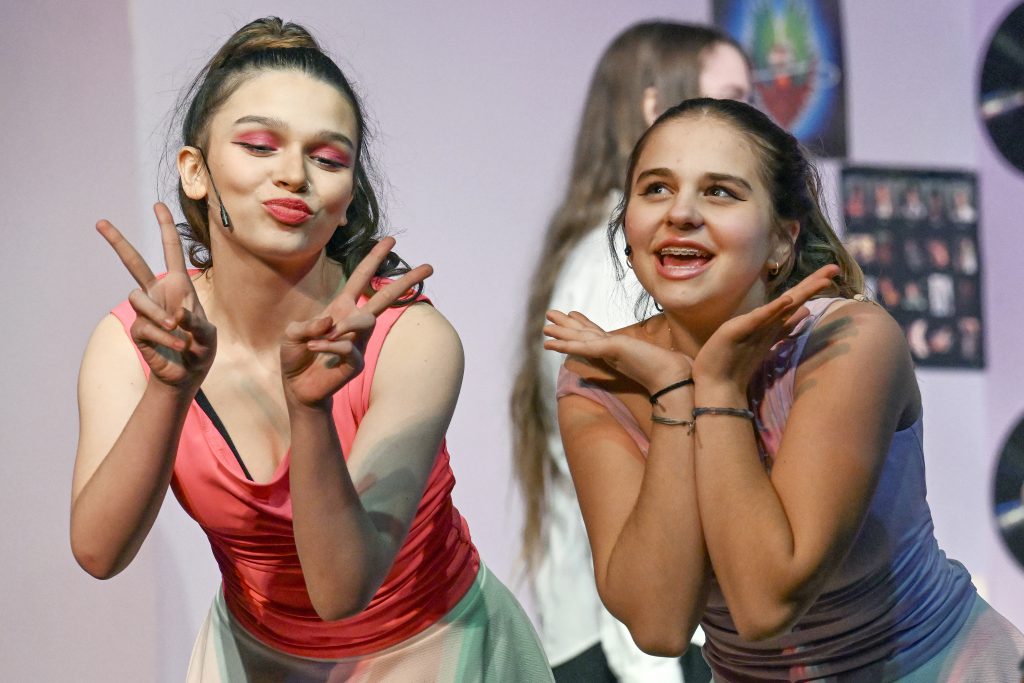 two girls wearing tan tops and pony tails sing a song together on the stage