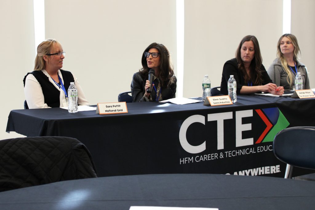 four adult women sit at a table with a tablecloth that reads "CTE," as one woman speaks into a microphone