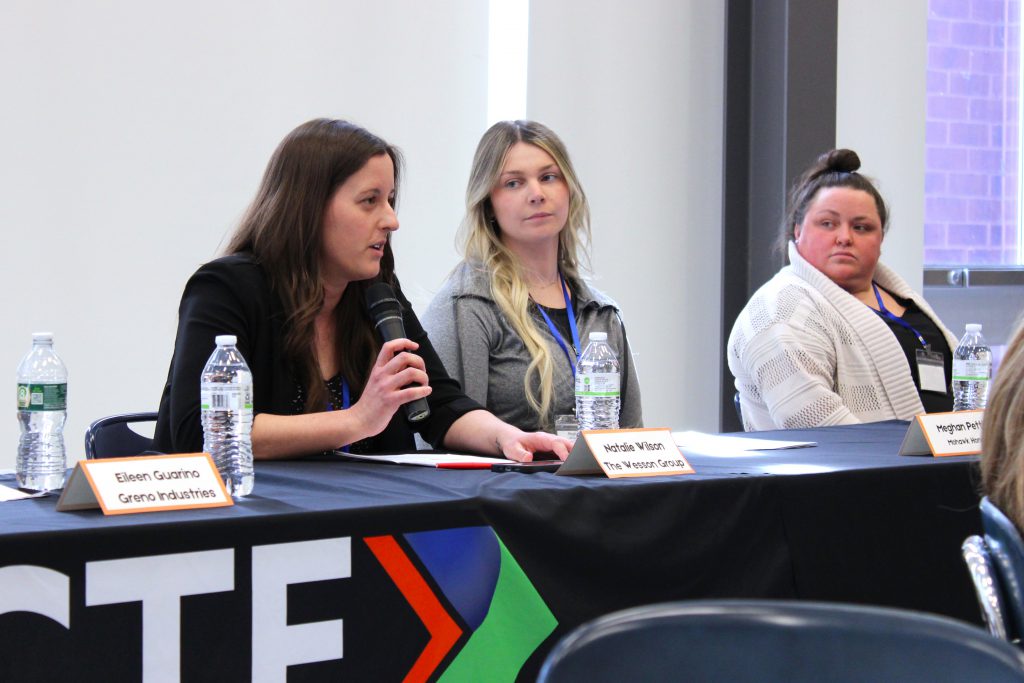 three women sit at a table with a black tablecloth, as one woman speaks into a microphone