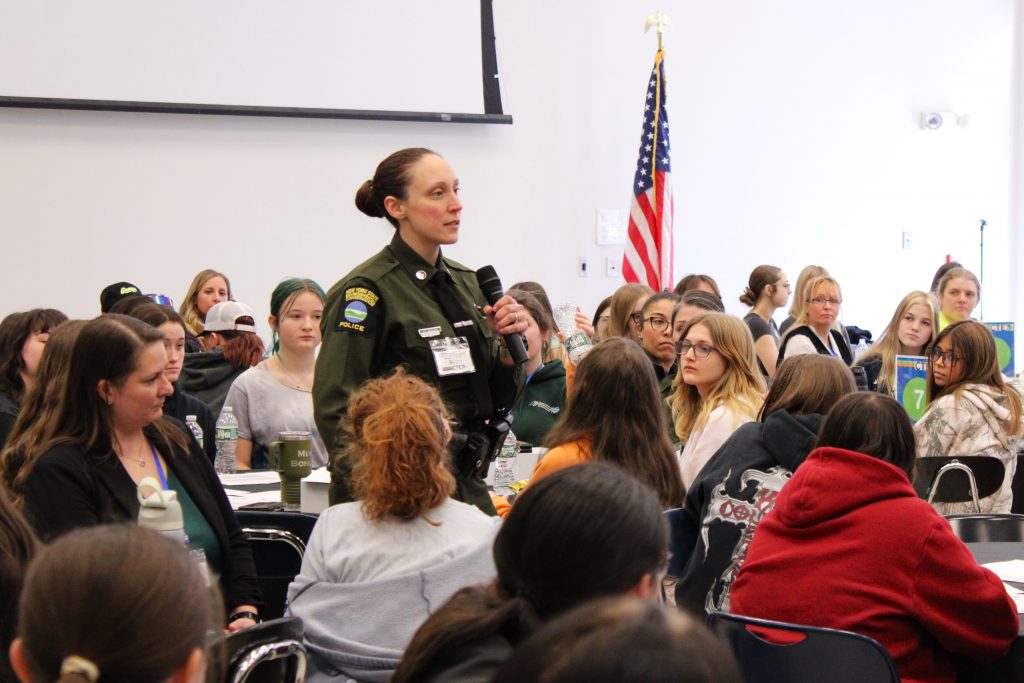 a female police officer speaks into a microphone as she stands in a room filled with young women 