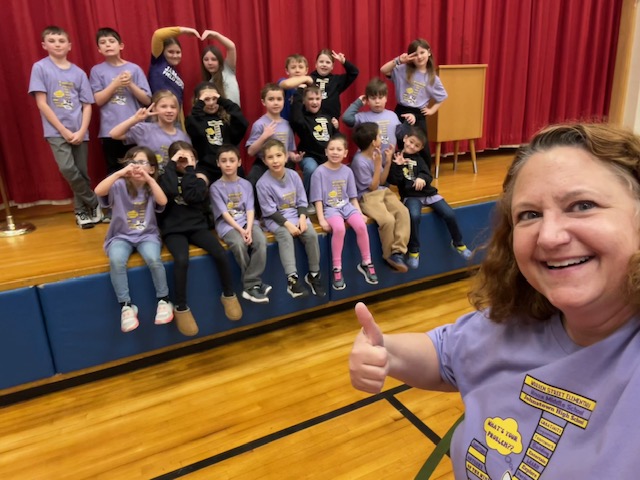 a teacher poses with a group of students behind her sitting on the stage