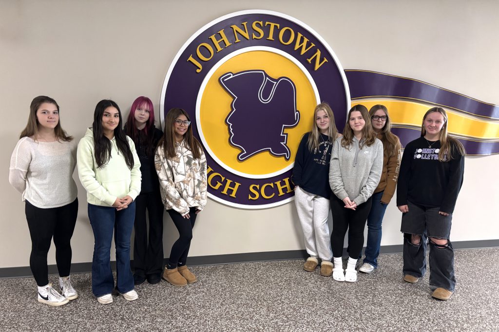 8 young ladies stand in the hallway in front of a purple and gold school logo that reads "Johnstown High School."