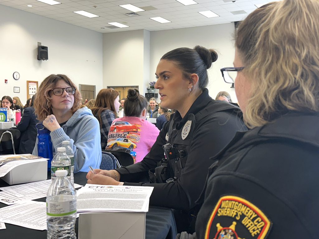 a police officer sits at a table and has a conversation with a colleague and a young female student